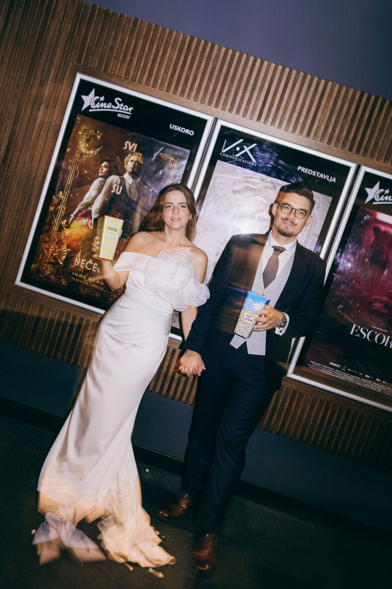 Mladenci se drže za ruke i poziraju u kinu ispred filmskih plakata. Mladenka u bijeloj vjenčanici s cvjetnim detaljem na dekolteu i mladoženja u elegantnom tamnom odijelu s naočalama drže kutije s kokicama.