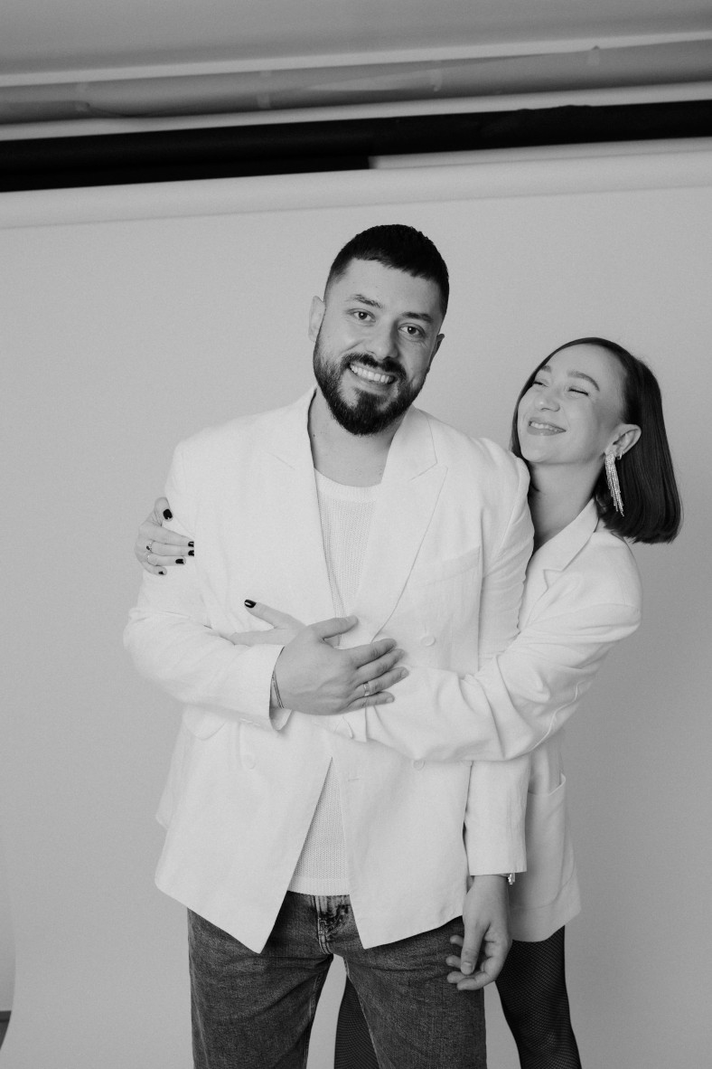 Radosna crno-bijela studijska fotografija muškarca s bradom i žene s kratkom kosom. Oboje nose bijele blejzere i smiju se dok žena grli muškarca s leđa. On gleda u kameru, a ona je nasmiješena. Pozadina je običan svijetli zid.