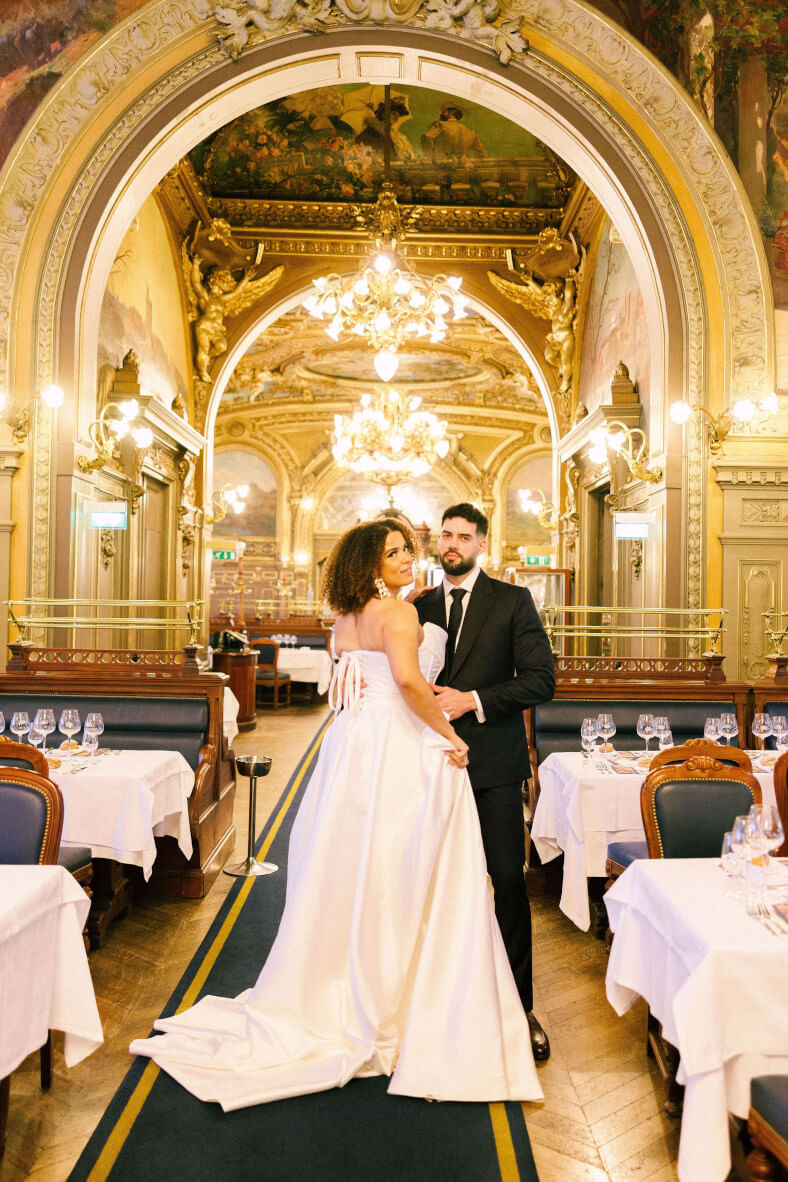 Mladenci poziraju u raskošno uređenom luksuznom restoranu sa zlatnim detaljima i kristalnim lusterima. Mladenka u elegantnoj bijeloj vjenčanici gleda prema kameri dok je mladoženja u crnom odijelu drži oko struka.