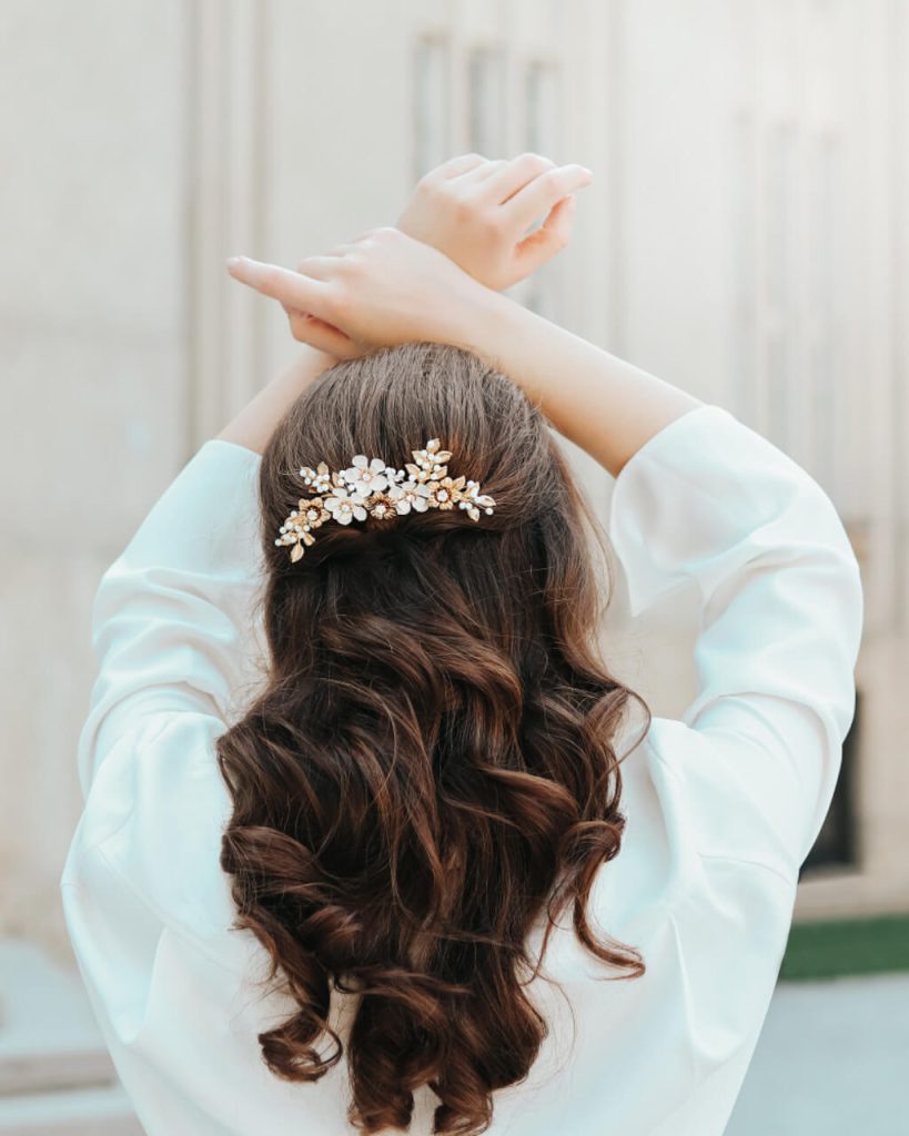 Pogled s leđa na ženu u bijeloj odjeći s podignutim rukama iznad glave. Njezina polupodignuta valovita smeđa kosa ukrašena je elegantnim zlatno-bijelim češljićem s cvjetnim motivima, biserima i sitnim kristalima.