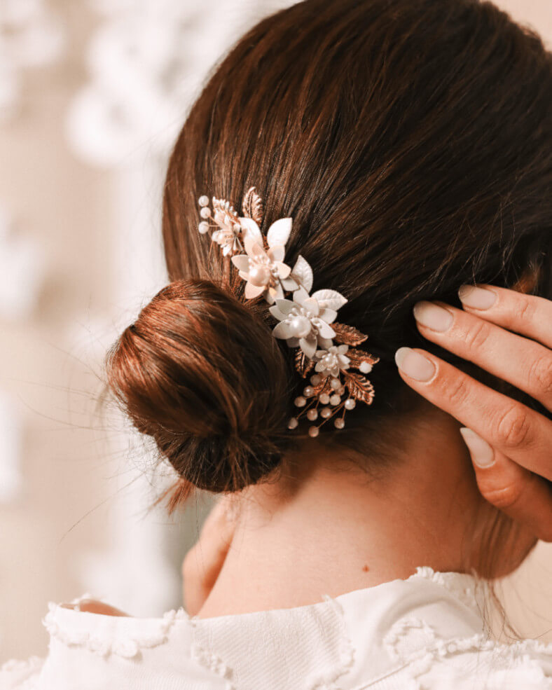 Pogled s leđa na elegantnu nisku pundžu ukrašenu bogatim floralnim ukrasom s bijelim cvjetićima, biserima i zlatno-bakrenim listićima, dok ruka nježno popravlja frizuru.