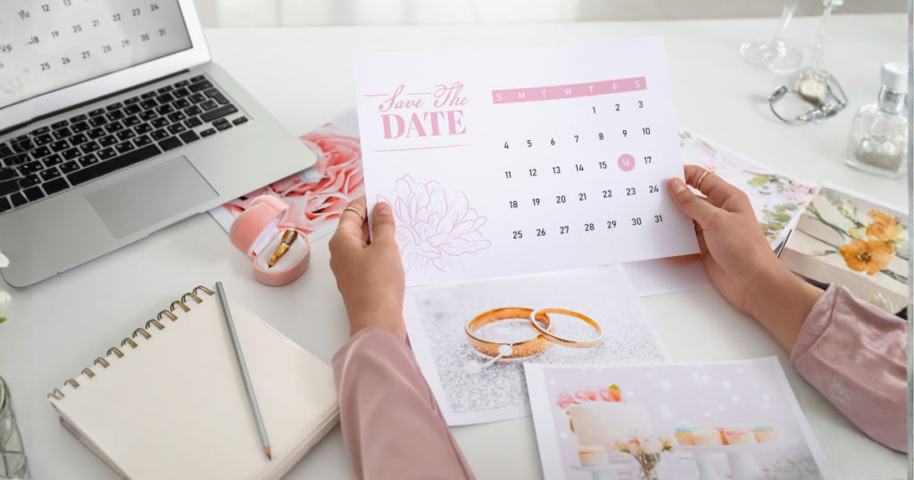 stol sa laptopom i raznim papirima te ruke koje drže papir na kojem je zaokružen datum i piše save the date (za vjenčanje)