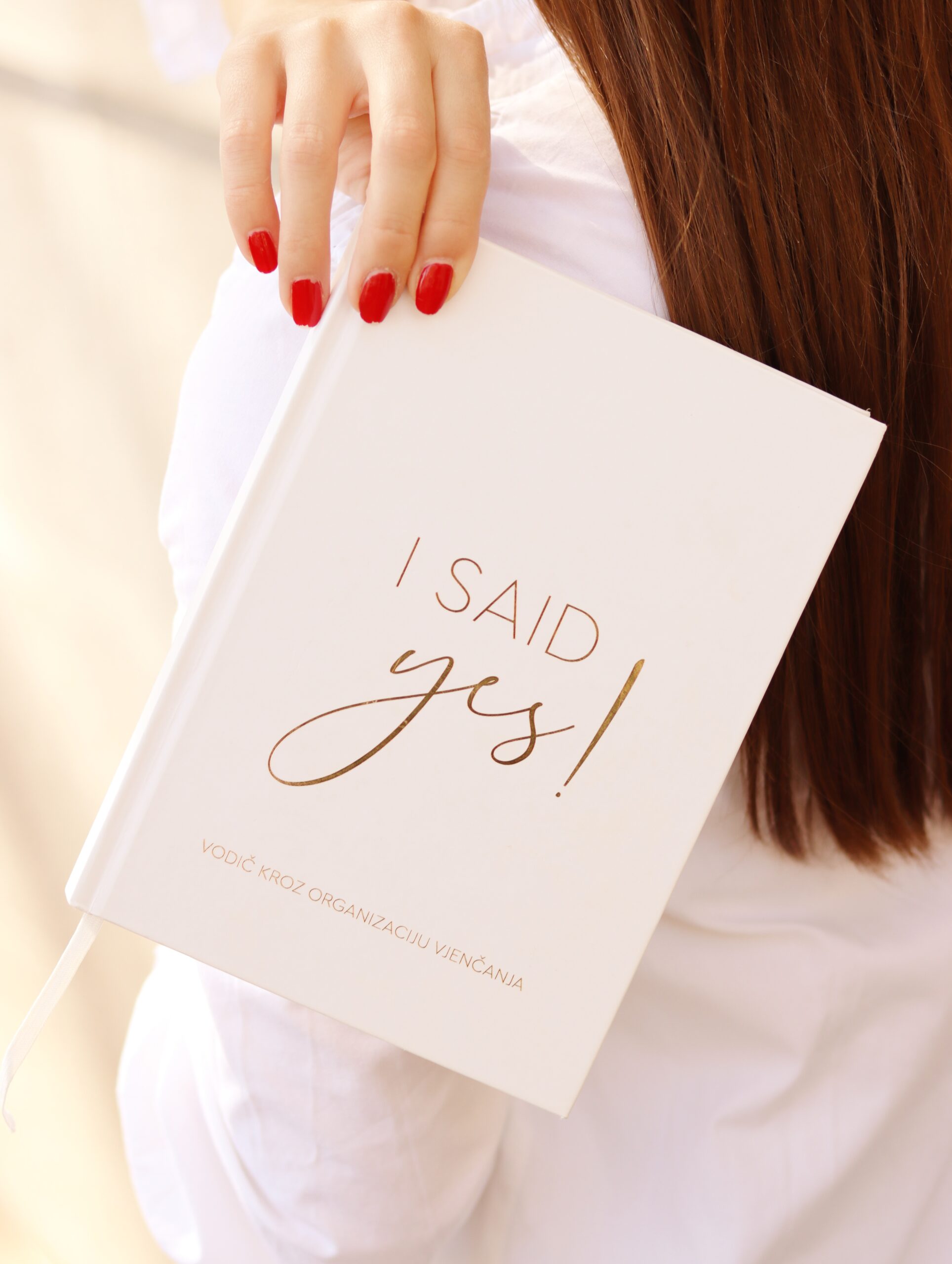Djevojka u bijeloj košulji drži bijeli I Said Yes planer vjenčanja sa zlatnim natpisom "I Said Yes! Vodič kroz organizaciju vjenčanja". Elegantni I Said Yes planer za vjenčanje – vodič za organizaciju vjenčanja i planer za mladenke.