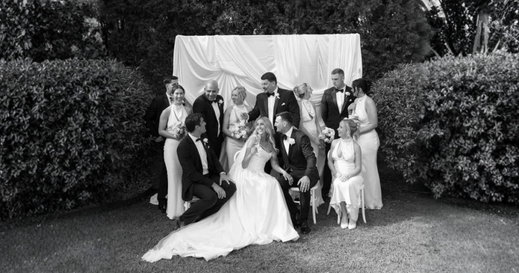 Crno-bijela grupna fotografija mladenaca i svatova u elegantnim odijelima i haljinama, koji poziraju u vrtu ispred bijele platnene pozadine okružene zelenilom