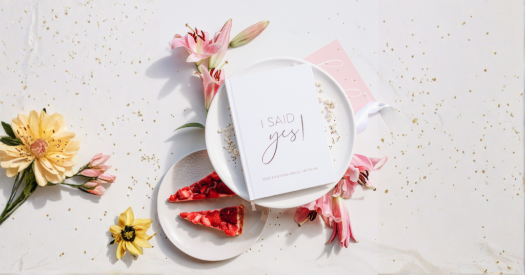 Pogled iz zraka na estetski aranžman s bijelim planerom za vjenčanje "I SAID yes!", ružičastim ljiljanima, žutim cvijećem, dva komada torte od jagoda na tanjuriću i posutim zlatnim šljokicama po bijeloj podlozi.