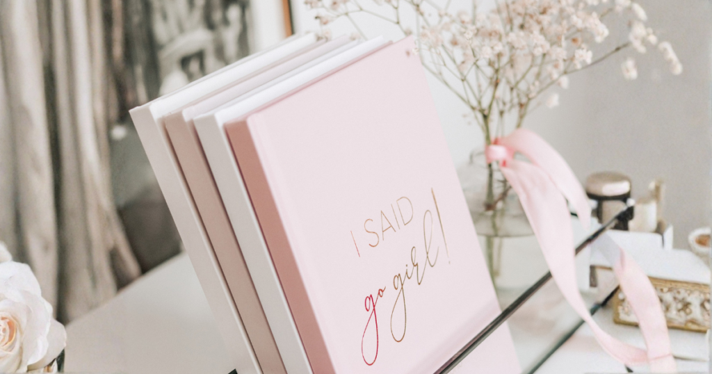 Složeni ružičasti i bijeli vjenčani planeri s natpisom "I SAID go girl!" izloženi na stalku, u okruženju staklene vaze sa gipsofilom i ružičastom trakom.